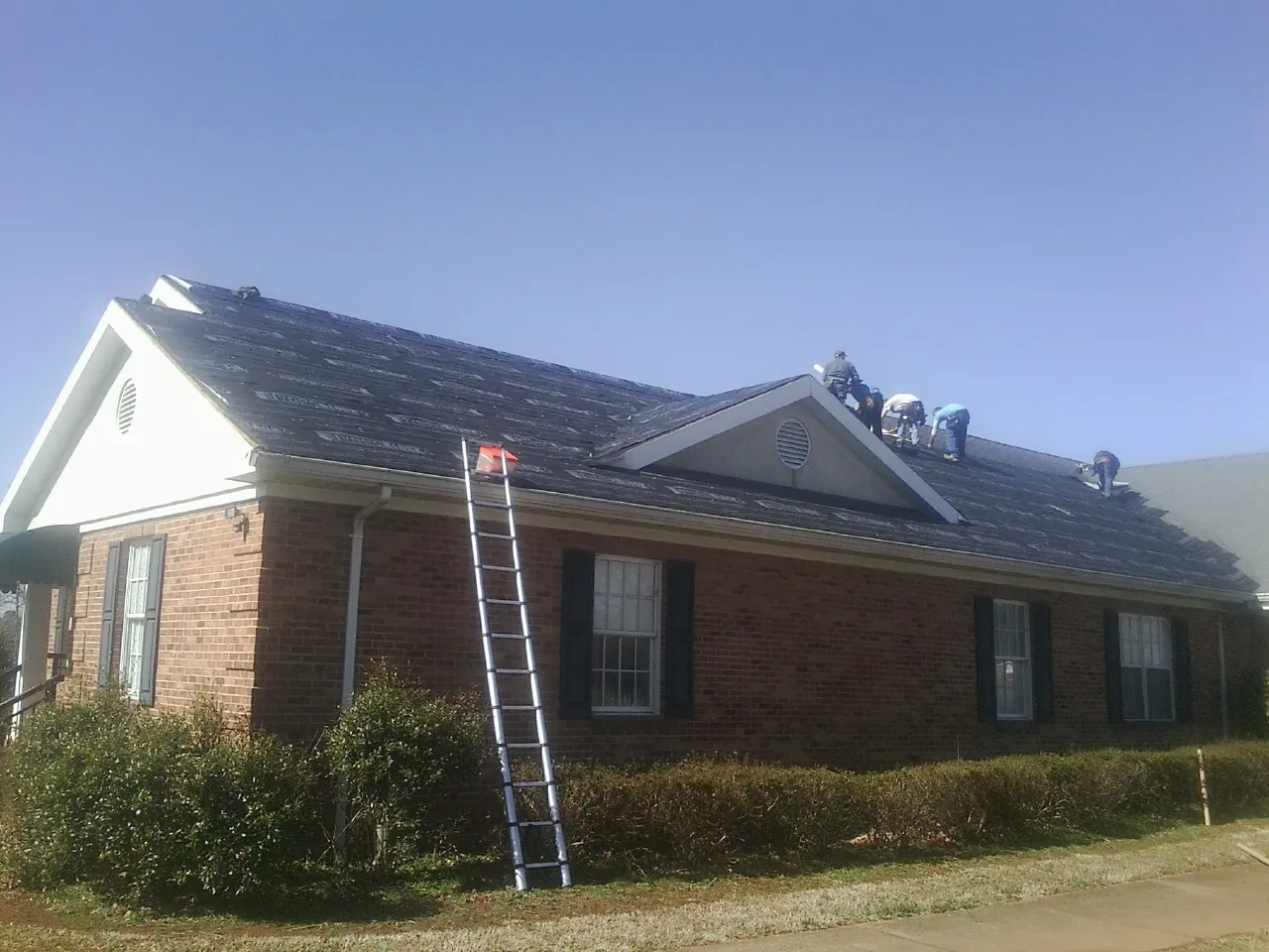 Crew tearing shingles off a brick ranch roof with a ladder set — Metal Roof Repair work by our crew in Judith Gap