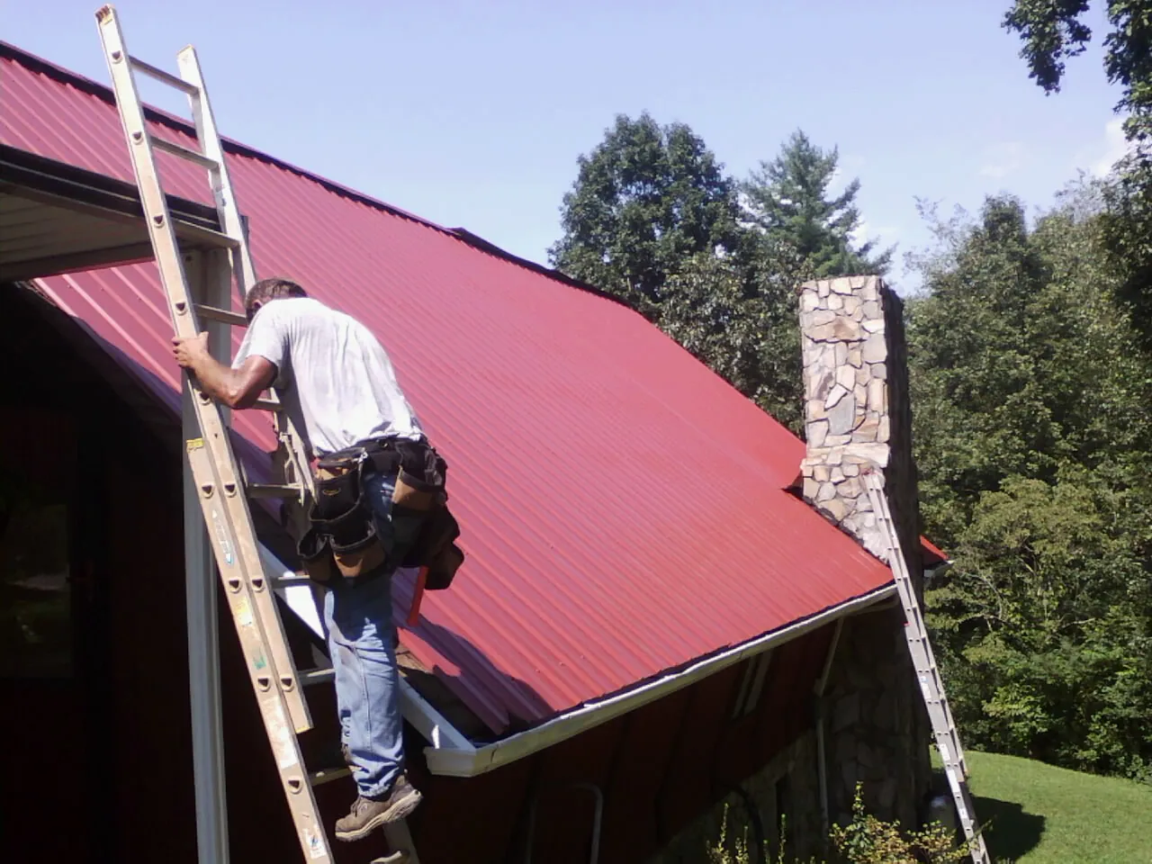 Roofer on a ladder beside a red metal roof and stone chimney — Soffit & Fascia Repair by Roofing Brothers in Judith Gap
